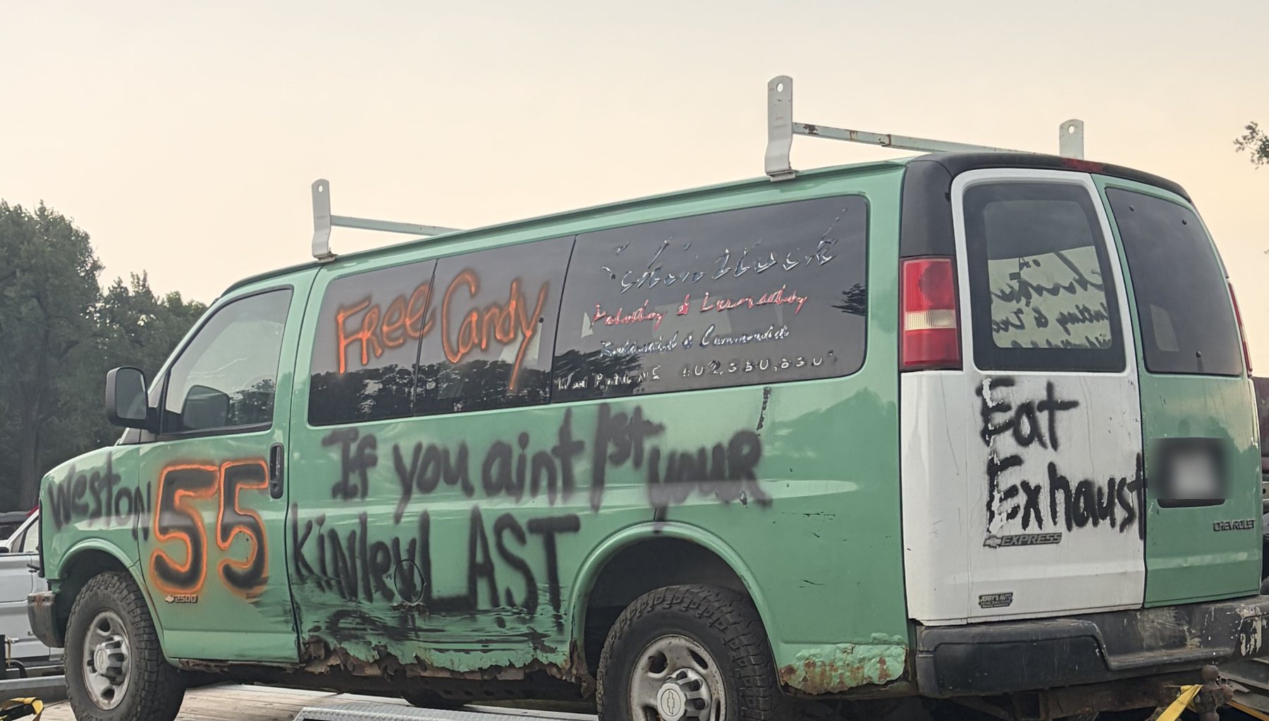 A mint green Chevrolet Express van loaded on a flatbed trailer at dusk, hand-painted with FREE CANDY in orange, a large number 55, the words 'If you ain't 1st you're last', and EAT EXHAUST across the rear doors.